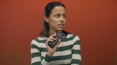 İspanyol kadın canlı kırmızı bir duvarın önünde uzaktan kumandayı tutuyor, dalgın ve odaklanmış görünüyor..