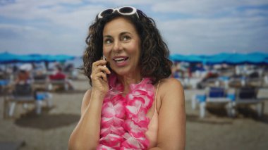 Güneşli bir sahilde telefonla konuşan bir kadın, bikini ve pembe bir lei giyiyor, güneş pansiyonları ve şemsiyelerle çevrili, deniz kenarındaki tropikal tatil havasının tadını çıkarıyor..