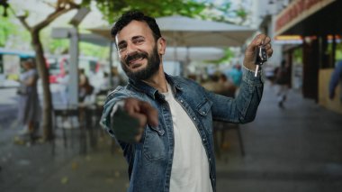 Sakallı İspanyol bir adam araba anahtarlarını güvenli bir şekilde bir sokak ortamında tutuyor, kot ceket giyiyor ve canlı bir şehir ortamında ağaçların altında gülümsüyor..