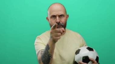 Futbol topu tutan sakallı kel bir adam, izole edilmiş yeşil arka plana karşı tehditkar hareketler yapıyor, duygularını ve yoğunluğunu açıkça ifade ediyor..