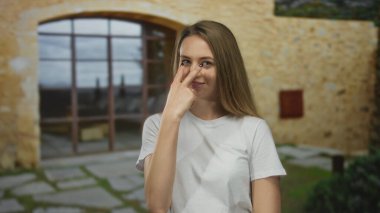 Açık havada, gözünün yanındaki parmağıyla el kol hareketi yapan, gözleri kapalı taş bir duvara poz veren, rahat bir gülümseme takınan, gözlem ve farkındalık hissi veren genç bir kadın..