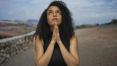 Açık havada dua eden kıvırcık saçlı, gözlüklü ve siyah gömlekli bir kadın bulutlu bir gökyüzünün altında sakin ve ruhani bir görünüm sergiliyor..