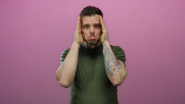 Dövmeli genç adam yüzünü ellerle kapatıyor pembe bir arka plana karşı üzüntüsünü ifade ederek güçlü bir duygusal görsel an yaratıyor..