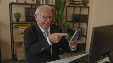 Akıllı telefon kullanan kıdemli bir adam modern ofiste masasında oturuyor ve başarılı haberlerin heyecanını ve neşesini gösteriyor.