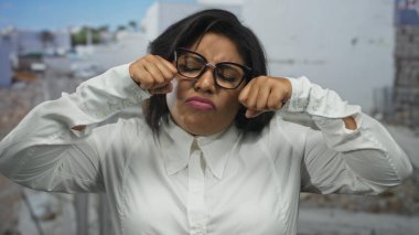 Gözlüklü genç bir kadın, bir inşaat alanında ağlama hareketi yapıyor, dışarıda duygularını ifade ediyor, Latin, artı beden, beyaz gömlek giyiyor..