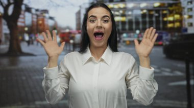 Kalabalık bir şehir caddesinde elleri havada gülümseyen genç İspanyol kadın, bulanık araba ve binalarla çevrili, şehir manzarasında sürpriz ve neşe ifade ediyor..