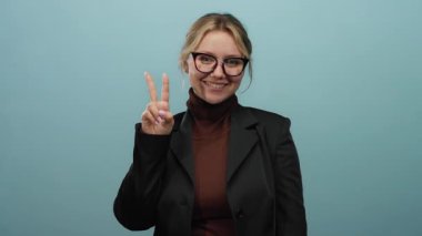 Siyah ceketli bir kadın mavi bir arkaplanda gülümsüyor neşeli bir ifadeyle barış işareti yapıyor ve gözlük takıyor..
