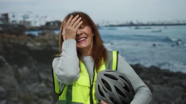 Deniz kenarında kahkaha atan, güvenlik yeleği giyen ve elinde kaskla okyanus dalgaları ve sakin bir günde arka planda kayalık bir plaj olan bir kadın..