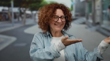 Middle-aged hispanic woman presenting with outstretched hands on a city street showing a joyful expression in an outdoor urban environment wearing glasses and a denim shirt.