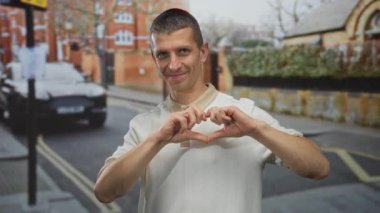 Sokaklarda dikilen bir adam elleriyle kalp şekli yaratıyor, binalar ve park halindeki arabalarla kentsel çevre tarafından geliştirilmiş sıradan bir ortamda mutluluk ve pozitiflik yayıyor..