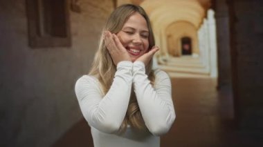 Üniversitenin manastır geçidinde kadın iki eliyle çerçeveler ve gözleri kapalı gülümsemeler; neşe oyunculuğu.