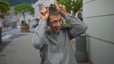 Güneşli bir şehir sokağında, arka planda ağaçlar ve mimari olan sıradan bir ortamda neşeli ve eğlenceli hareketler yapan bir adam..