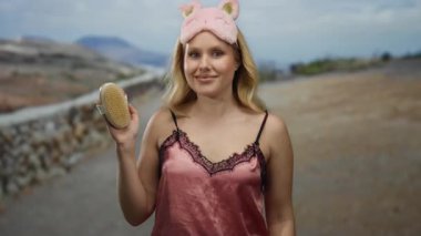 Gece elbiseli kadın, sakin dağ ortamında fırça tutuyor, uyku maskesi takıyor, doğa ile çevrili, sakin sabah rahatlığı ve doğal güzelliği somutlaştırıyor..