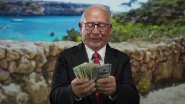 Dolarları olan kıdemli bir adam takım elbisesiyle deniz kenarında gezinti yapıyor..