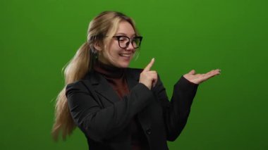 Gözlüklü ve siyah ceketli gülümseyen sarışın kadın, boş bir alana işaret ediyor. Canlı yeşil izole arka plana karşı. Boş bir konsept jest ve sunum sergiliyor.