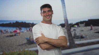 Gülümseyen adam kollarını kavuşturup kumlu sahil sahilinde okyanus ufkunun yakınındaki bir palmiye ağacının altında çıplak kollarını gösteriyor..