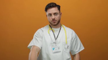 Sakallı ve steteskoplu genç bir doktor izole edilmiş turuncu arka planda kameraya doğru bakıyor. Tıbbi bağlamda güven ve profesyonellik yayıyor..