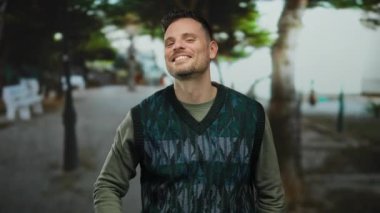 Şehir sokaklarında gülümseyen ve kameraya bakan genç adam şehir ağaçlarının altında neşeli yüz ifadesini sergiliyor..