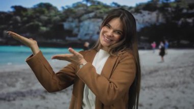 Genç İspanyol kadın, iki eliyle sahilin manzaralı sahnesinde turkuaz deniz ve arka planda bulanık ağaçlarla bir şey sunuyor..