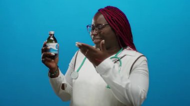 Beyaz bluzlu gülümseyen kadın bir ilaç şişesi gösteriyor ve mavi arka planda, gözlüklü ve boynunda steteskop olan el kol hareketi yapıyor..
