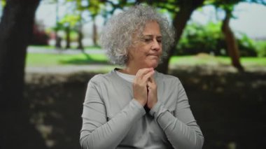 Gri saçlı, güneşli bir park ortamında dalgın dalgın duran yaşlı bir kadın..