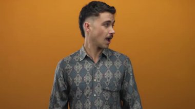 Şablonlu tişört giyen genç İspanyol adam canlı turuncu bir arka plana şaşırmış görünüyor, izole bir ortamda merak ve duygu yayıyor..