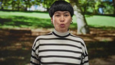 Genç bir kadın açık hava parkında, yemyeşil ağaçların arasında komik bir yüz ifadesi sergiliyor, neşeli ifadeler sergiliyor ve doğal bir çevrede enerjik bir enerji sergiliyor..