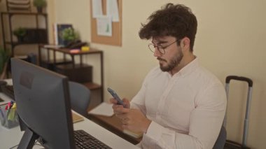 Sakallı genç adam ofiste bilgisayar masası ve kapalı alandaki bitkilerle selfie çekiyor. Modern teknoloji ve mobilyaların yer aldığı rahat bir profesyonel atmosfer yaratıyor.