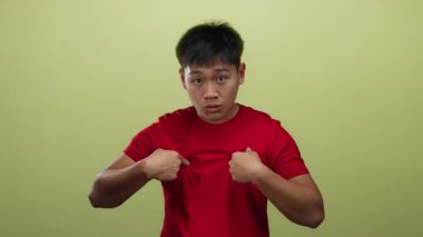 Kırmızı tişörtlü genç Çinli adam, izole edilmiş sarı arka plan duvarına karşı kuşkulu bir ifadeyle kendini işaret ederek, mizahi ve karışık bir hava yaratıyor..