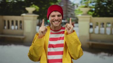 Sarı ceketli ve kırmızı bereli genç adam, şehir sokaklarında gülümsüyor, neşeli ve neşeli bir hava sergiliyor, bulanık şehir manzarası var..