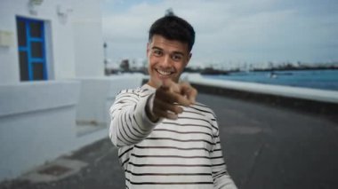 Deniz kenarında bir gezinti güvertesinde gülümseyen genç adam çizgili bir gömlek giyiyor ve neşeli bir ifade takınıyor, manzaralı bir deniz manzarası ile çevrili..