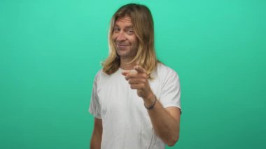 Beyaz gömlekli, uzun sarı saçlı, gülümseyen adam yeşil stüdyoda bana jest yapıyor..