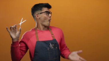 Gözlüklü genç İspanyol adam elinde makas tutarak canlı turuncu arka planda, saç şekillendirmedeki eğlence ve yaratıcılığı ifade ediyor..