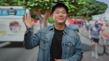 Kot ceketli genç Çinli adam ağaçlar ve yayalar kameraya el sallıyor..