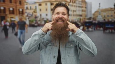 Uzun sakallı bir adam işlek bir caddede şehir binaları ve geçen kalabalıklar arasında parmaklarını dişlerine işaret ediyor..