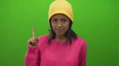 Pembe kazaklı ve sarı bereli İspanyol kadın izole edilmiş yeşil bir duvara karşı ciddi bir ifadeyle tetikte bir jest yapıyor..