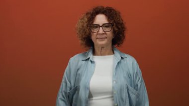 Orta yaşlı esmer, esmer ve gözlüklü, esmer, esmer bir kadın kollarını düz kahverengi bir duvara yaslamış, sıradan bir kot gömlek giyiyor..