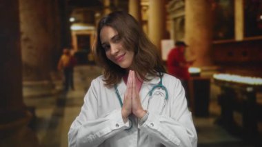 Steteskoplu genç İspanyol kadın doktor kilisede duruyor, ellerini birleştirip dua ediyor, profesyonel kıyafetleri ve ruhani ayarı kutsal bir mekanda harmanlıyor..