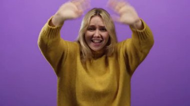 Canlı mor bir arka plana karşı ellerini kaldırmış duygularını ifade eden bir kadın, sarı saçlı ve sarı kazaklı, izole ve dinamik bir anı yakalıyor..