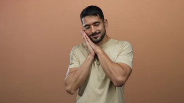 Genç adam kahverengi arka planda dinleniyor, elleri çenesinin altında ve gözleri kapalı, sakinlik ve sükunet yayıyor, açık bir gömlek giyiyor ve huzurlu bir ifade sergiliyor..