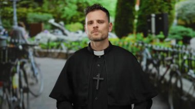 Yeşillik bir park ortamında dikilen papaz resmi bir ifade ve dini kıyafetle, arka planda bulanık yapraklarla çevrili..