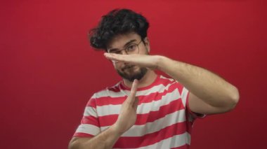 Sakallı ve gözlüklü genç adam, parlak kırmızı bir arkaplanda, çizgili gömlek giyiyor, el hareketleriyle duraksama ya da kırılma belirtileri gösteriyor, İspanyol kültürünü sergiliyor..