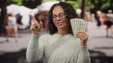 Orta yaşlı bir kadın şehir sokaklarında Amerikan dolarlarını elinde tutuyor. Kendine güvenen ve gülümseyen bir jest yapıyor..