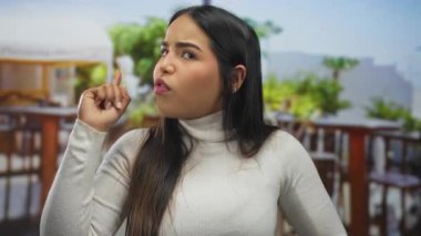 Kadın, restoranın içindeki bir sunucuyu işaret ederken parmağını kaldırıyor. Genç Latin kökenli bir kadın terasta dikiliyor ve kızgın görünüyor..