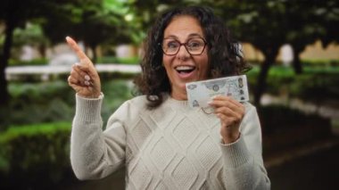 Parkta orta yaşlı bir kadın elinde sürpriz bir ifadeyle Dirham banknotunu tutuyor. Heyecan ya da keşif öneriyor..