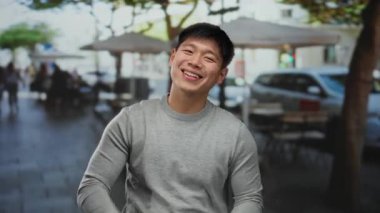 Genç Asyalı adam güneşli bir restoranın terasında otururken neşeli bir şekilde gülümsüyor ve arka planda insanlar var..