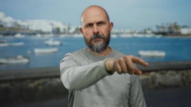 Deniz kenarında sakallı bir adam arka planda görünen teknelerle açık gökyüzünün altında gri bir gömlek giyiyor ve sert bir ifade sergiliyor..
