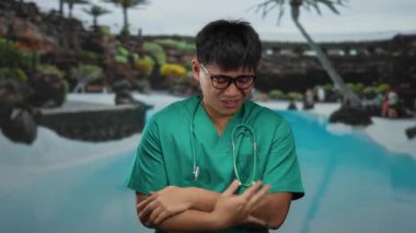 Yeşil üniformalı genç adam açık hava otelinde serin bir günde havuz kenarında üşümüş görünüyor..