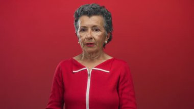 Gri saçlı yaşlı bir kadın canlı kırmızı bir arkaplanda endişeli görünüyor, duyguları ve yaşı, gözleri kapalı ve elleri alnında yakalıyor..