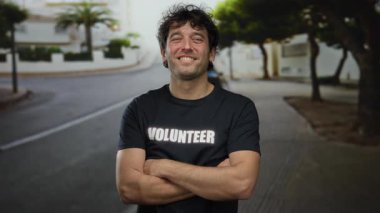 Gönüllü tişört giyen İspanyol adam, güneşli caddede ağaçlar ve binalarla kaplı çapraz kollarla kendinden emin bir şekilde gülümsüyor, açık havada toplum ruhunu somutlaştırıyor..